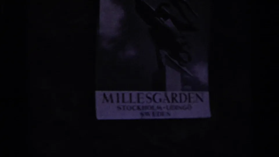 Nazare home-movie frame showing a dim sign or poster area with the readable word “MILLESGARDEN” beneath a shadowed sculptural form.
