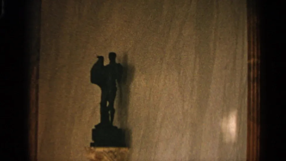 In 1965 Nazaré home-movie footage, a small dark standing figurine appears on a pedestal against a warm tan interior wall.