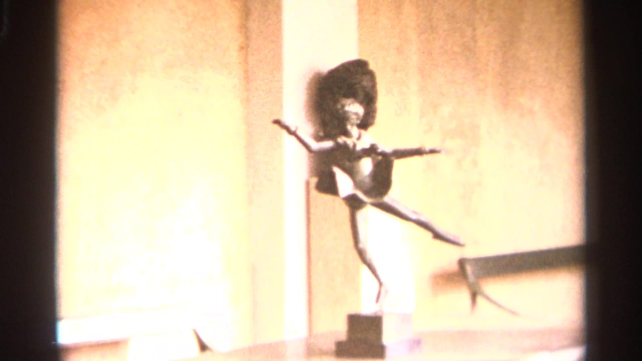 A small dancer-like sculpture on a pedestal is filmed indoors in Nazaré, Portugal, with bright 8mm exposure on the wall behind it.