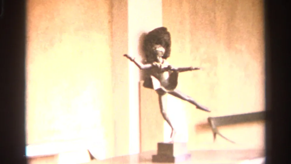 A small dancer-like sculpture on a pedestal is filmed indoors in Nazaré, Portugal, with bright 8mm exposure on the wall behind it.
