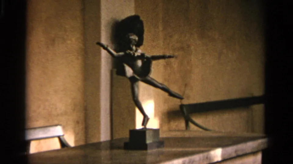 The 1965 8mm frame shows a dancer sculpture on a plinth, with a wall light, tan plaster surface, and interior ledge visible nearby.