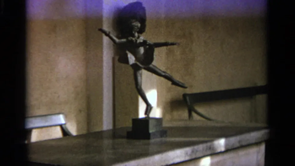 A handheld 8mm view centers the dancer figurine on an interior ledge in Nazaré, Portugal, with warm walls and a small light fixture.