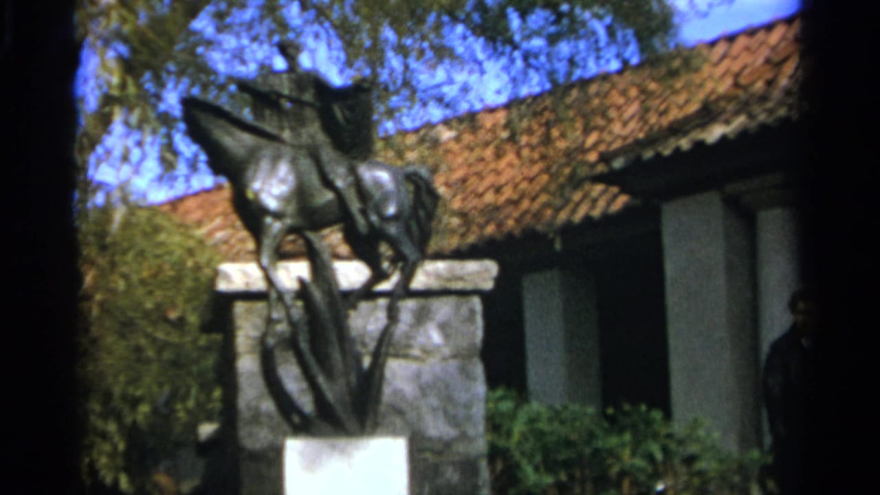 1965 Nazaré, Portugal 8mm frame showing a bronze soldier-on-horse statue on a stone pedestal near a tiled-roof building.