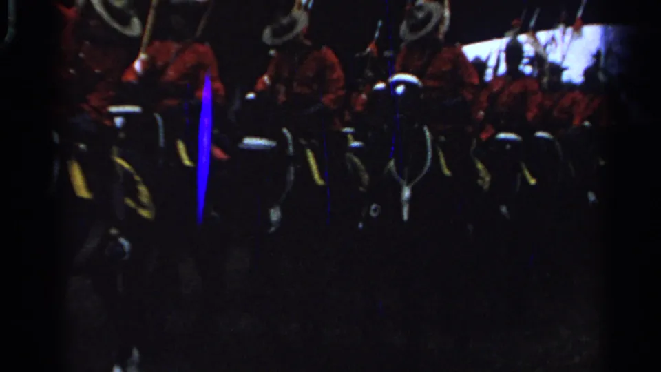 A close, dark 8mm view shows red-uniformed horseback riders with white helmets and vertical blue film scratches.