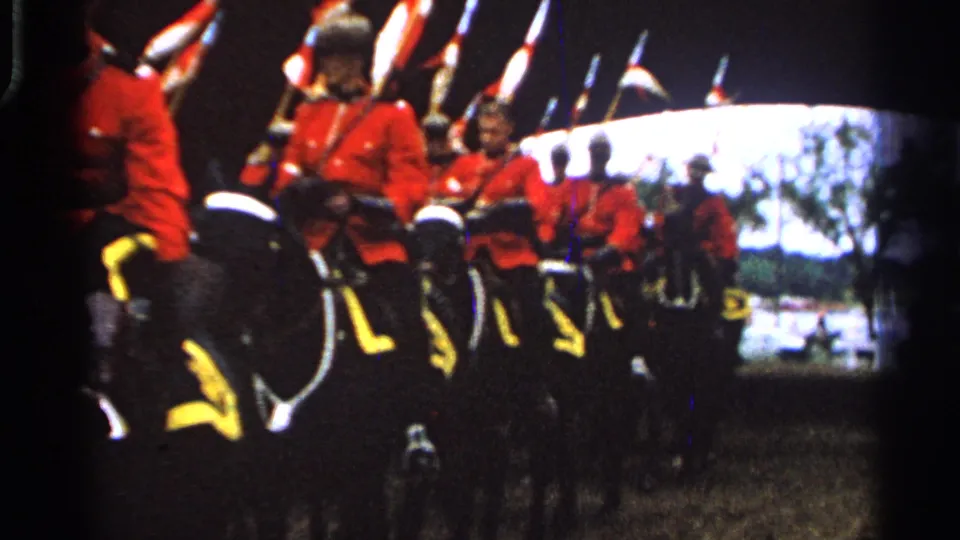 The procession continues as mounted riders in red jackets and dark hats pass through the frame with flags rising above the formation.