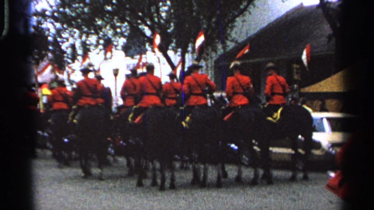 Mounted officers in red coats ride black horses along a street with spectators and trees nearby.