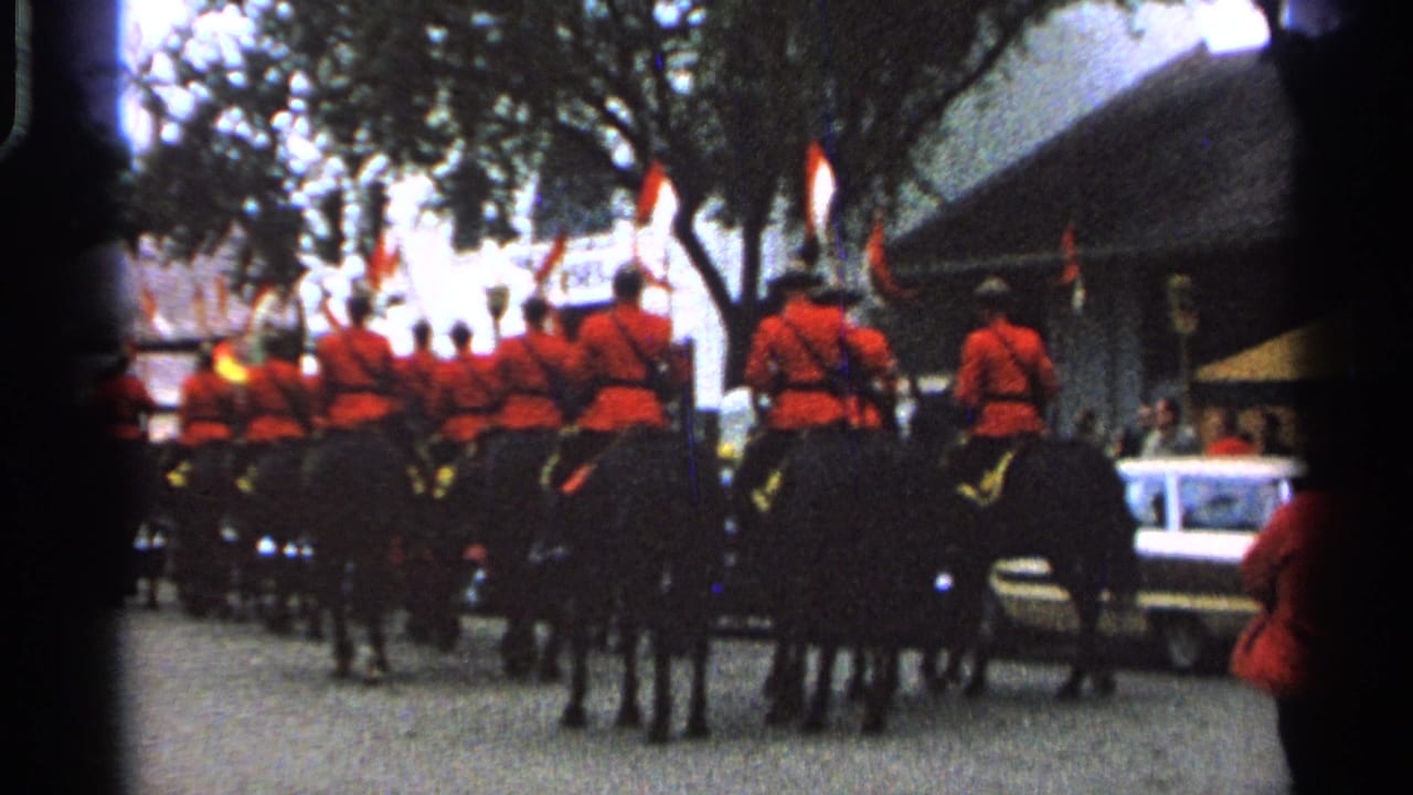 Mounted riders in red uniforms continue down the street beside trees, spectators, and cars.