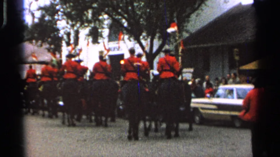 1960s parade view with mounted police in red uniforms, dark horses, roadside spectators, trees, and a light-colored sedan in Saint Louis.