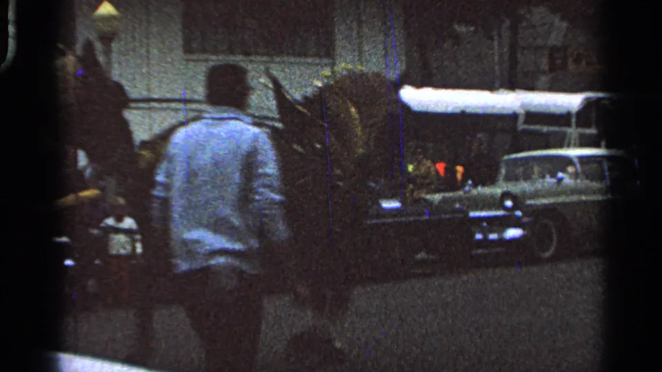 Street-level Saint Louis parade frame showing a bystander near horses, a covered vehicle, and a classic car as the mounted procession passes.