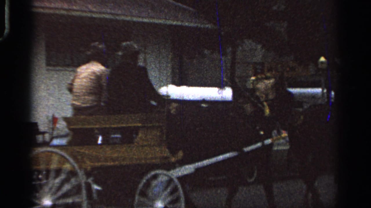 Two men in hats ride or sit in a wooden horse-drawn buggy during a nighttime 1965 Saint Louis state fair scene.