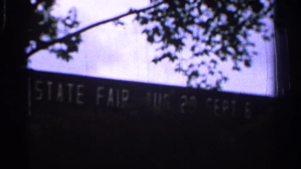 A dark 8mm frame shows a sign reading STATE FAIR beneath tree branches, supporting the fairground setting in the 1965 Missouri footage.
