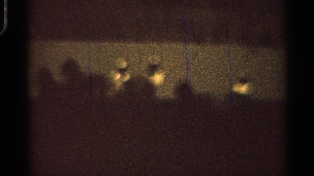 Low-light 8mm view of a nighttime fair event, with small bright lights and indistinct figures near the horizon.