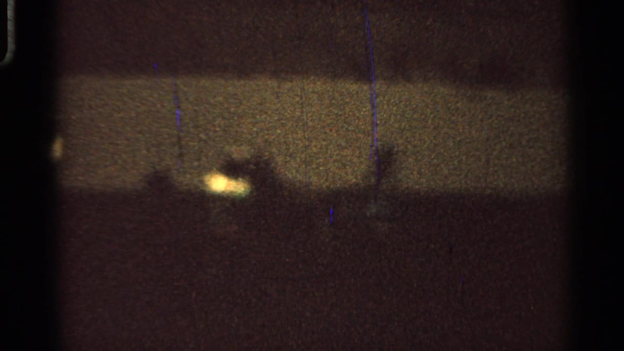 Grainy 1965 Missouri fairground frame showing distant silhouettes and bright points of light in a dark outdoor arena.