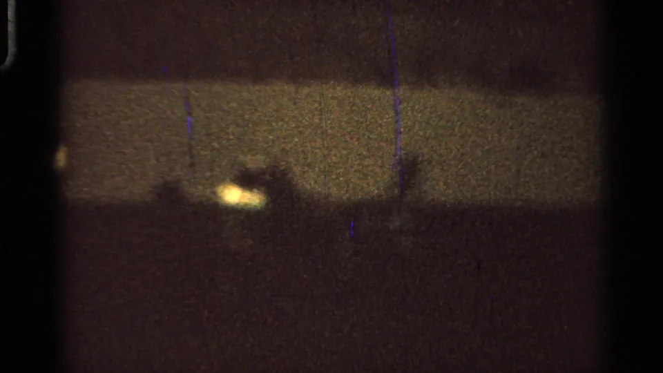 Grainy 1965 Missouri fairground frame showing distant silhouettes and bright points of light in a dark outdoor arena.