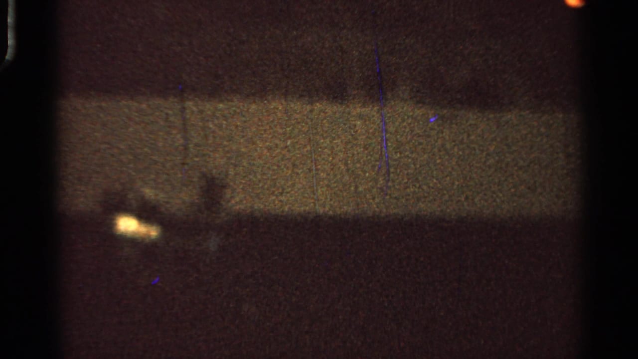 1965 Saint Louis 8mm frame with dim silhouettes, a small yellow light, heavy grain, blue scratches, and dark frame borders.
