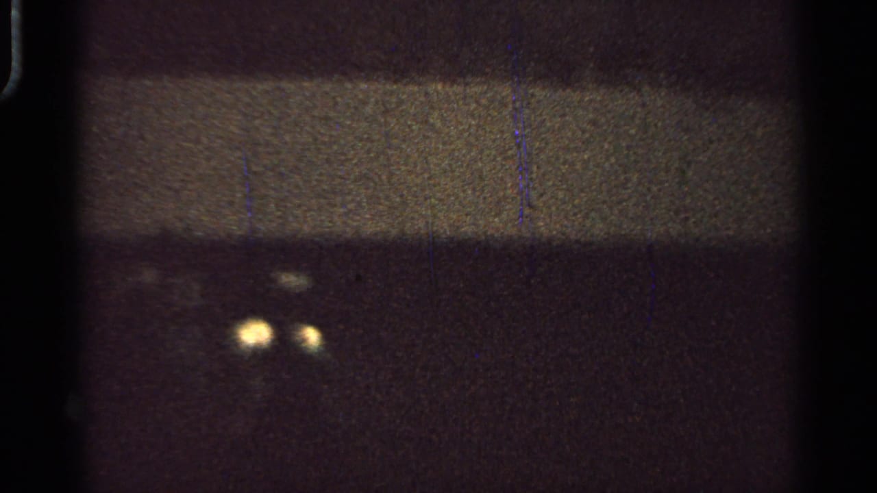 1965 Saint Louis 8mm frame showing two faint yellow lights against a dark, grainy field with scratches and black film edges.