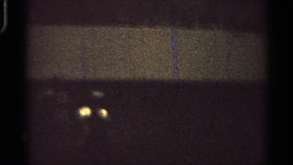 1965 Saint Louis 8mm frame showing two faint yellow lights against a dark, grainy field with scratches and black film edges.
