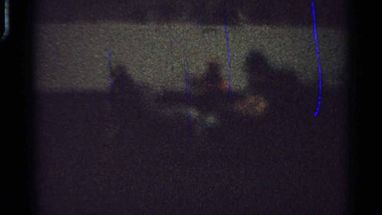 Low-light 1965 Saint Louis frame with shadowy figures and a probable carriage or horse shape crossing an indoor event floor behind a barrier.