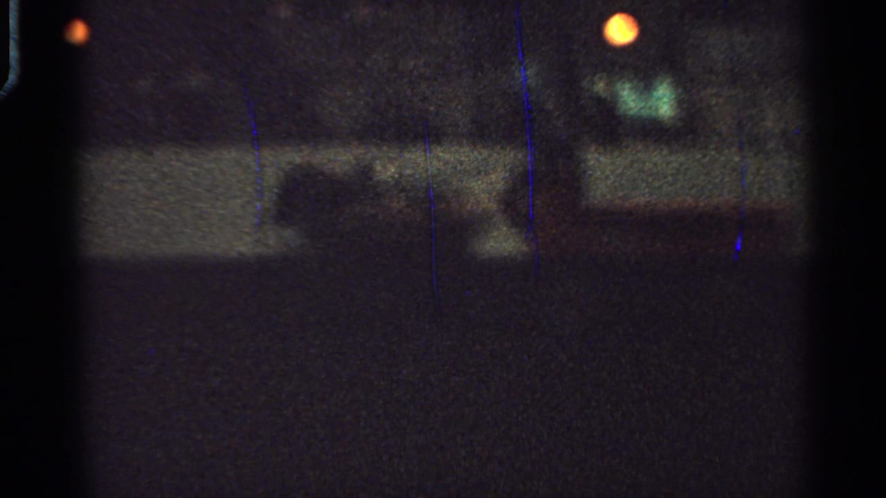 Grainy 1965 Saint Louis 8mm frame showing an indoor event space with warm light, greenish illumination, barrier rails, and indistinct people.