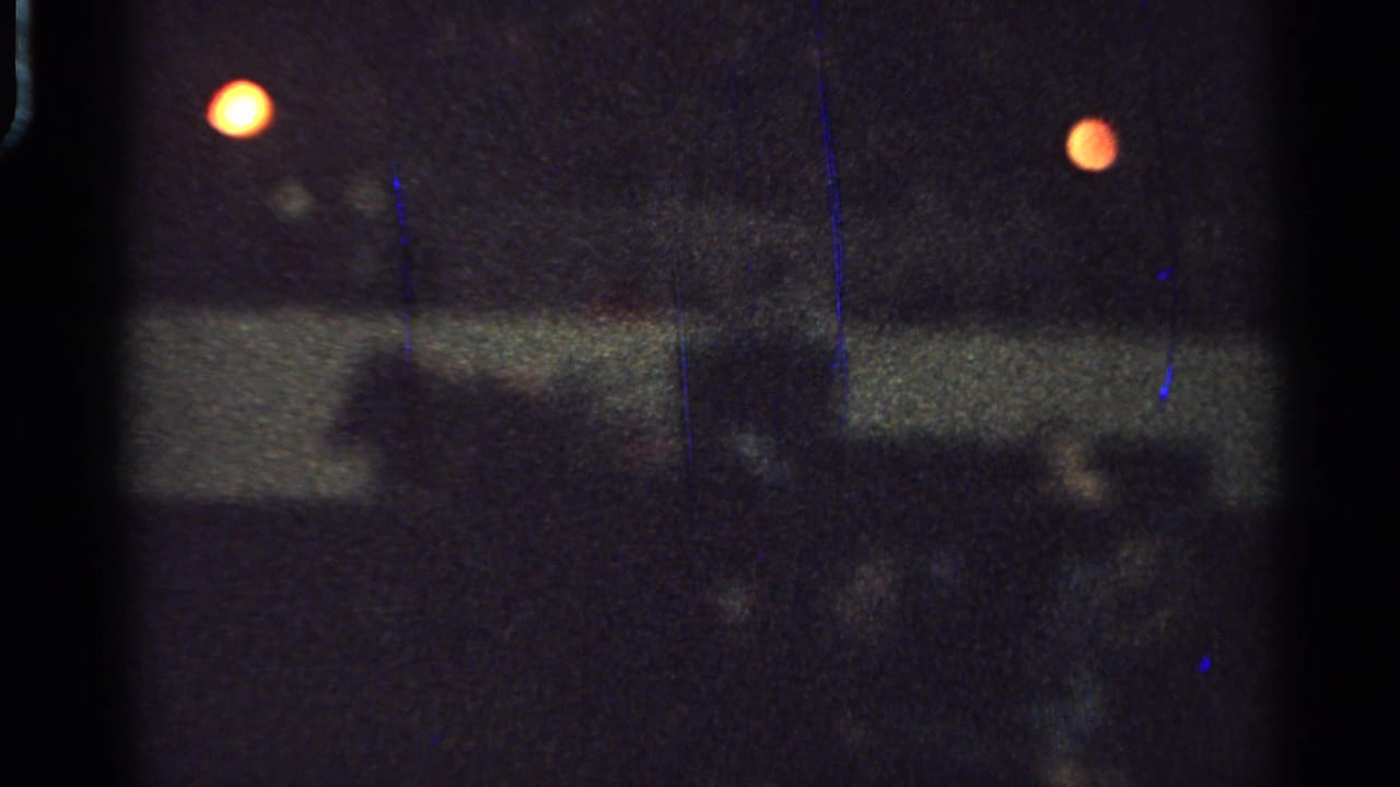 Soft 1965 Saint Louis home-movie frame of a dark indoor event area with rail barrier, warm lights, and blurred spectator silhouettes.
