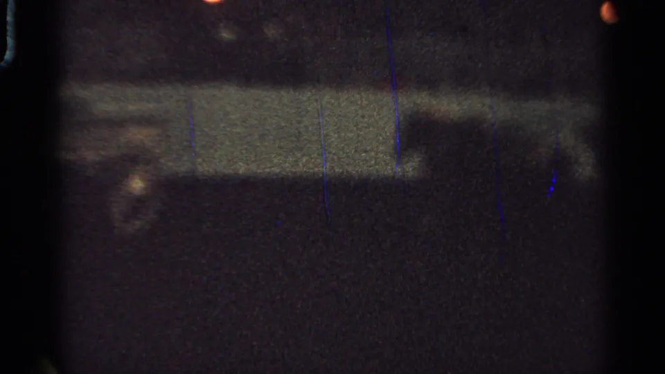 Low-exposure 1965 Saint Louis 8mm frame showing a pale barrier-like band, an orange light, and indistinct motion in a dark arena setting.