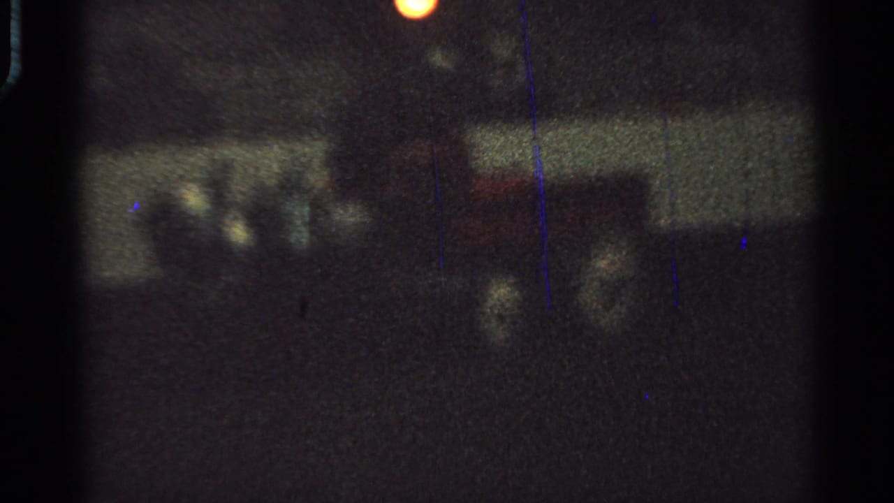 Blurry 1965 Saint Louis 8mm frame with indistinct figures or cart forms in a dark nighttime setting and a small warm light flare.