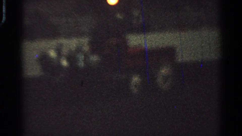 Blurry 1965 Saint Louis 8mm frame with indistinct figures or cart forms in a dark nighttime setting and a small warm light flare.