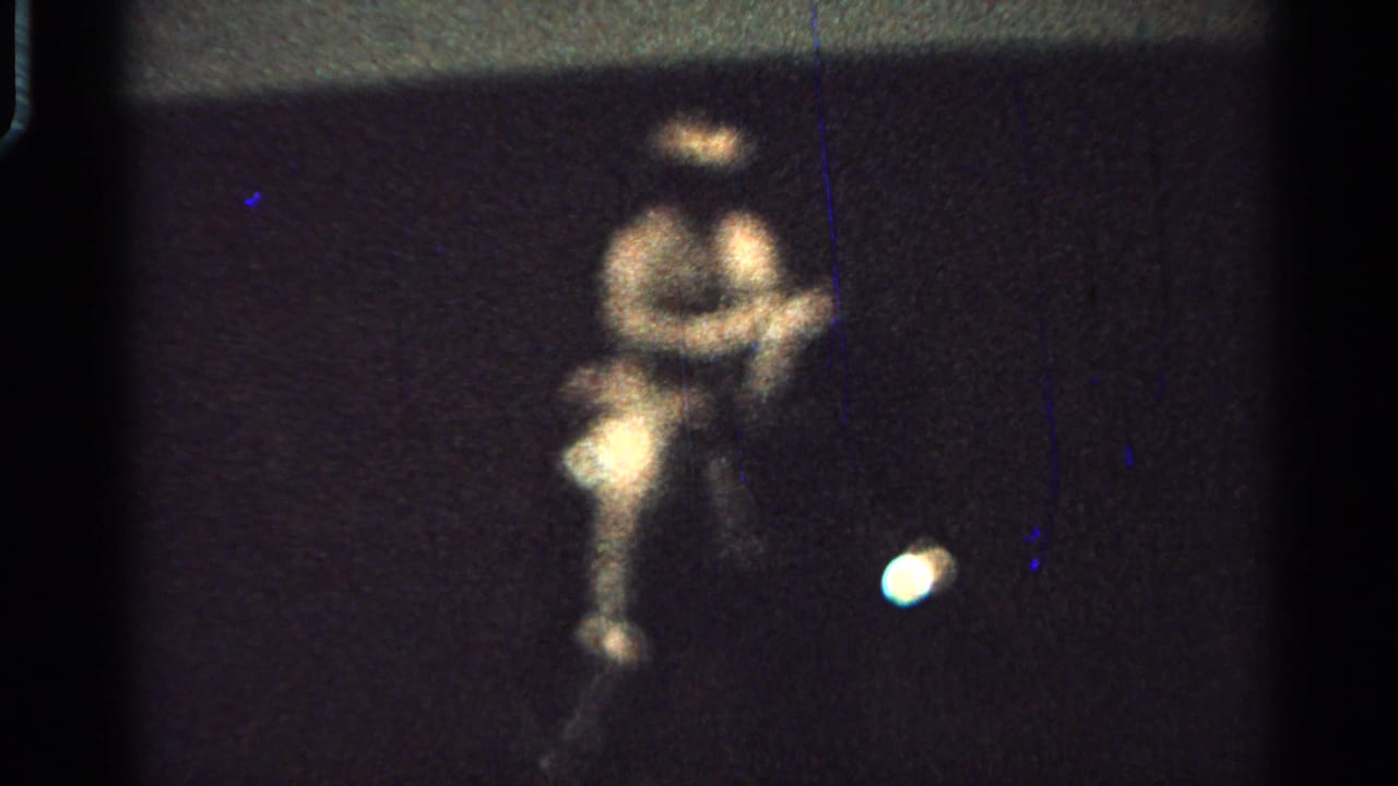 The 8mm frame shows a closer blurred view of the seated rider on a dark cart, with grain, scratches, and a bright spot near the lower right.