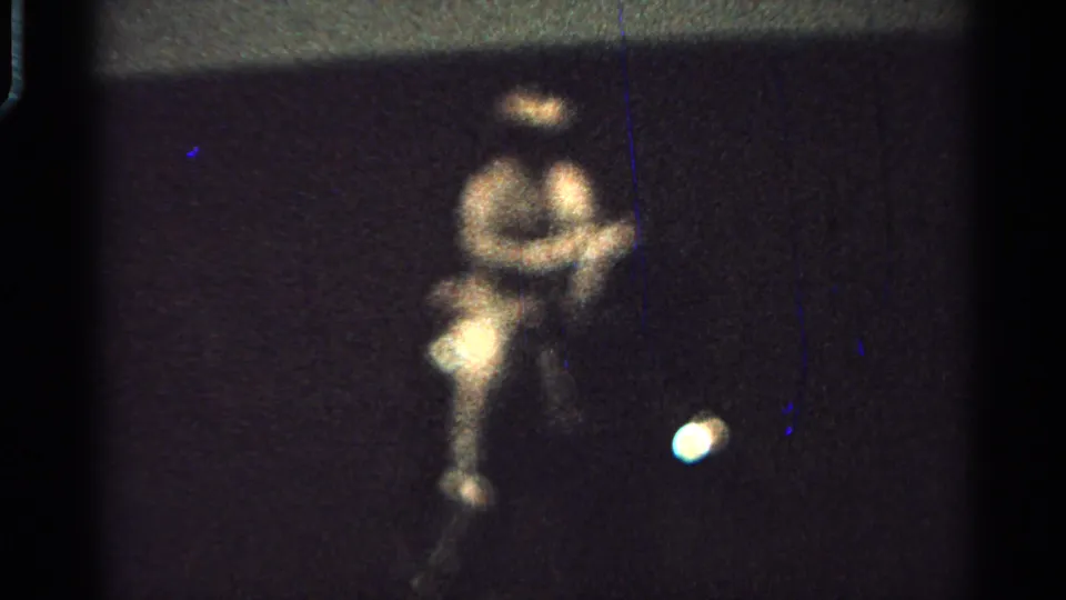 The 8mm frame shows a closer blurred view of the seated rider on a dark cart, with grain, scratches, and a bright spot near the lower right.