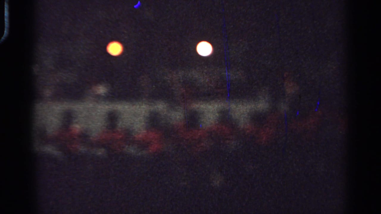 Dark grainy frame with red-clad figures below two bright circular lights.