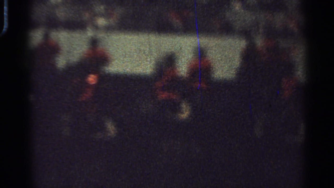Dark mounted riders in red coats pass along a pale barrier in low light.