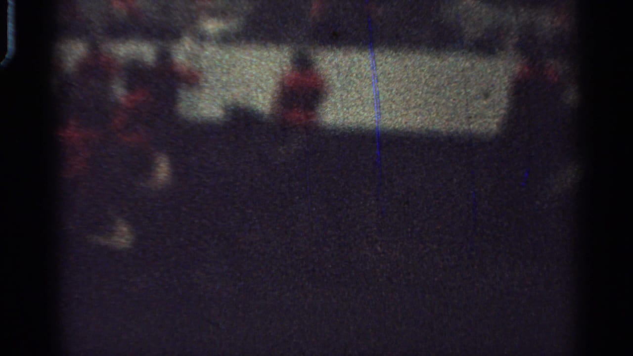 Soft 1965 Saint Louis home-movie frame showing a dim parade view with red-clad riders or attendants near an arena barrier.