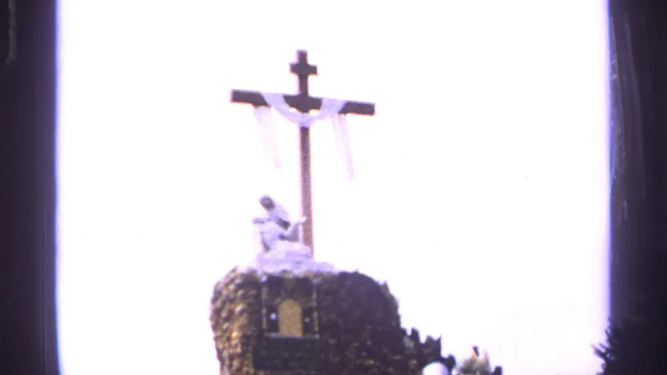 Saint Louis 1965 home-movie frame showing a crucifix with a small statue below it, silhouetted against bright sky.
