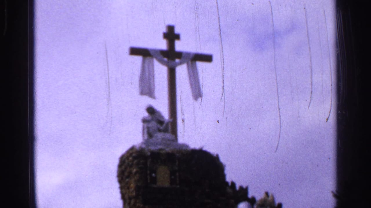 1965 Saint Louis 8mm frame showing a wooden cross draped with white cloth above a small religious statue on rough stone.