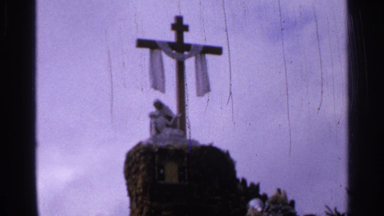 Soft 8mm view of a draped cross and small statue on a rocky monument, with pale sky behind it in Saint Louis metadata context.