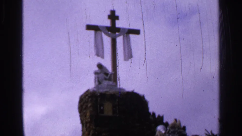 Soft 8mm view of a draped cross and small statue on a rocky monument, with pale sky behind it in Saint Louis metadata context.