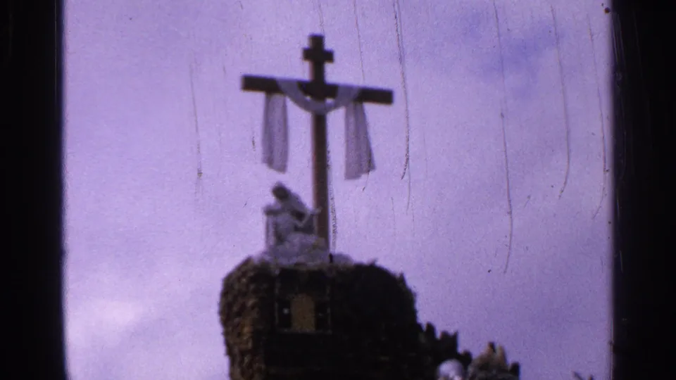 Saint Louis 1965 home-movie frame of a draped wooden cross above a small statue, softened by faded color and film wear.