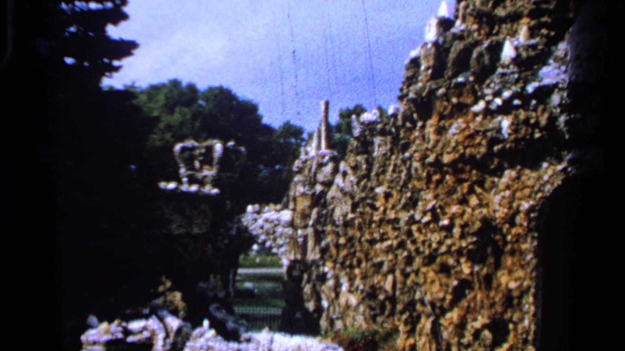 1965 Saint Louis frame with castle-like stone garden elements, dense tree shade, and a glimpse of open garden beyond.