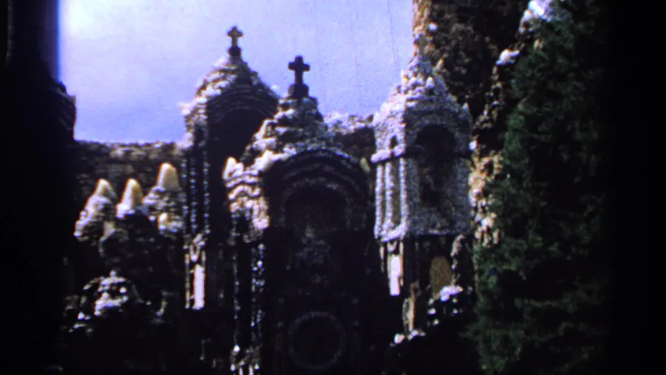 1965 Saint Louis 8mm frame showing a stone religious shrine with arched niches, crosses, dense carvings, and trees at the edge of the scene.