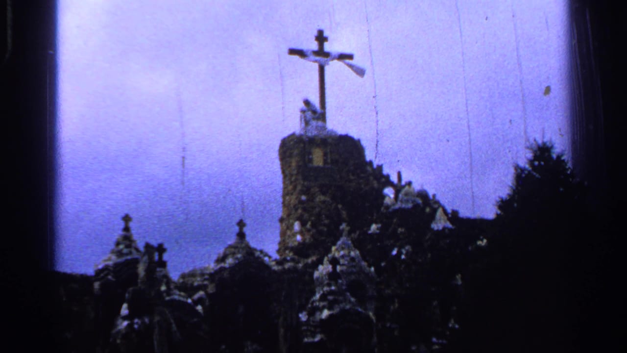 Tall cross on a stone tower above smaller shrine rooftops and dark trees.