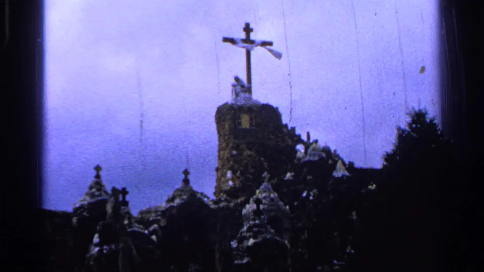 1965 Saint Louis 8mm frame with a tall cross rising from a stone tower, smaller shrine structures below, and trees silhouetted at the right.