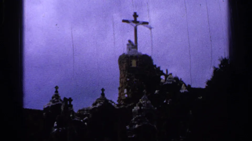 1965 Saint Louis 8mm frame of a tall cross on a stone tower, with shadowed shrine rooftops below and trees against a purple-tinged sky.