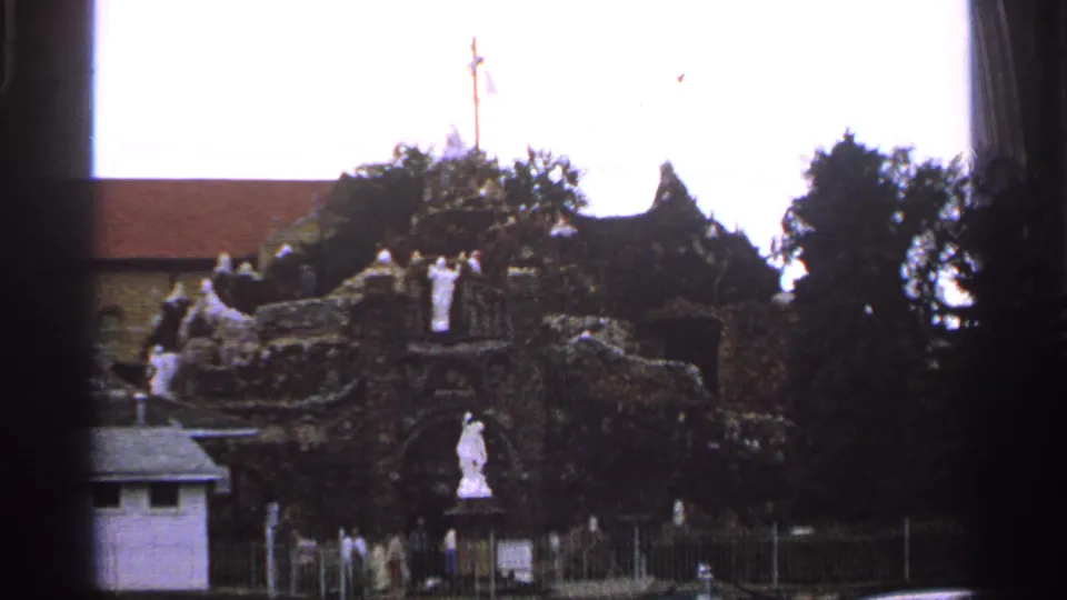 Wider 1965 Saint Louis frame showing a stone religious monument with statues, a cross, trees, fencing, and distant visitors.