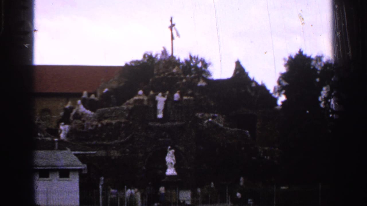 1965 Saint Louis home-movie frame showing a distant shrine or grotto with white statues, a cross, trees, and dark foreground framing.