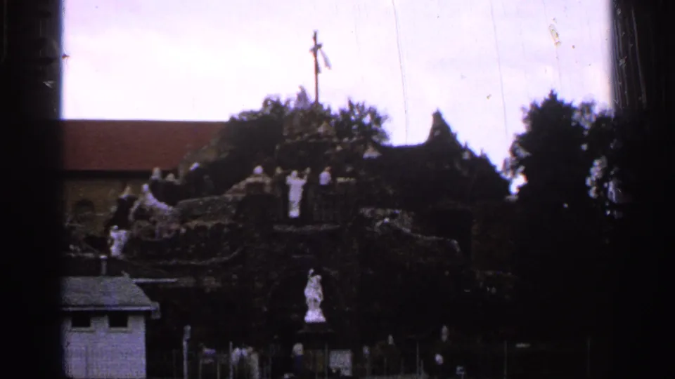 1965 Saint Louis home-movie frame showing a distant shrine or grotto with white statues, a cross, trees, and dark foreground framing.