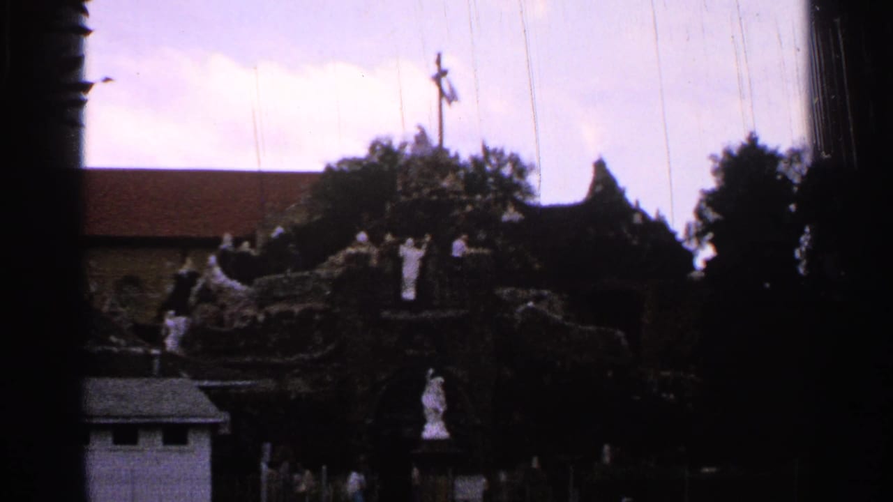 Religious shrine with statues on a rocky structure and a cross rising behind trees.
