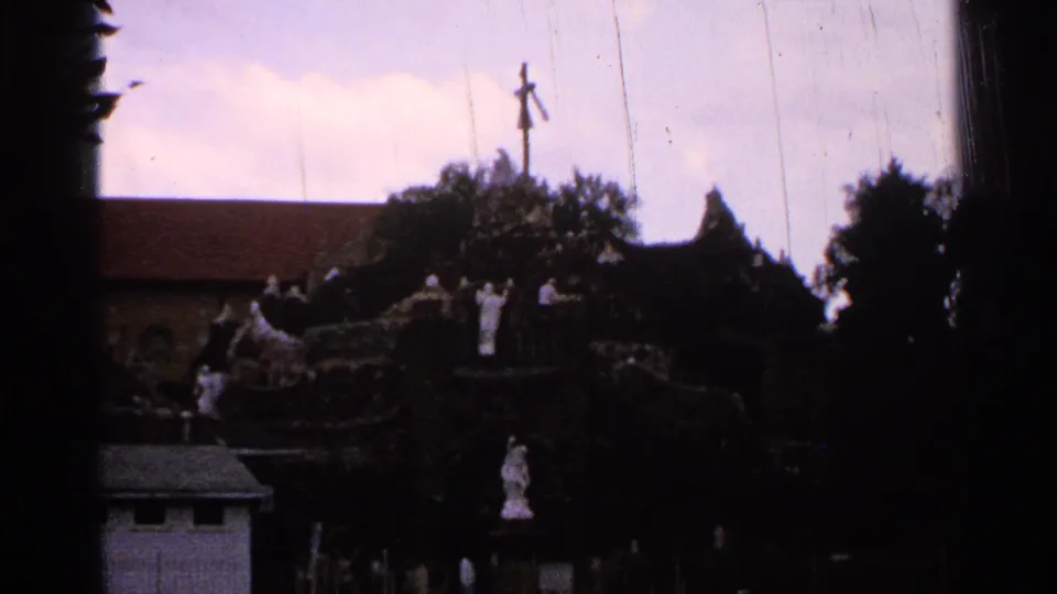 1965 Saint Louis home-movie view of a religious grotto seen from a distance, with saint statues, a cross, trees, and building roofs nearby.