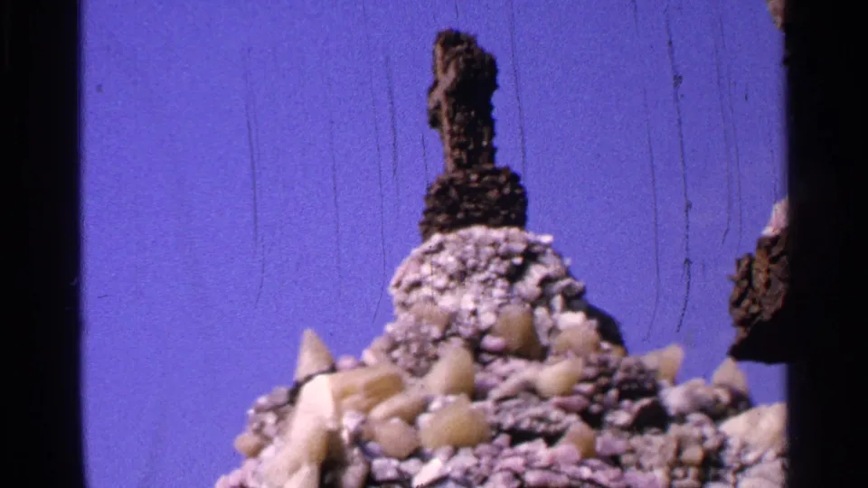 1965 Saint Louis home-movie frame showing a rocky Easter display with a dark cross-like sculpture against a blue painted backdrop.