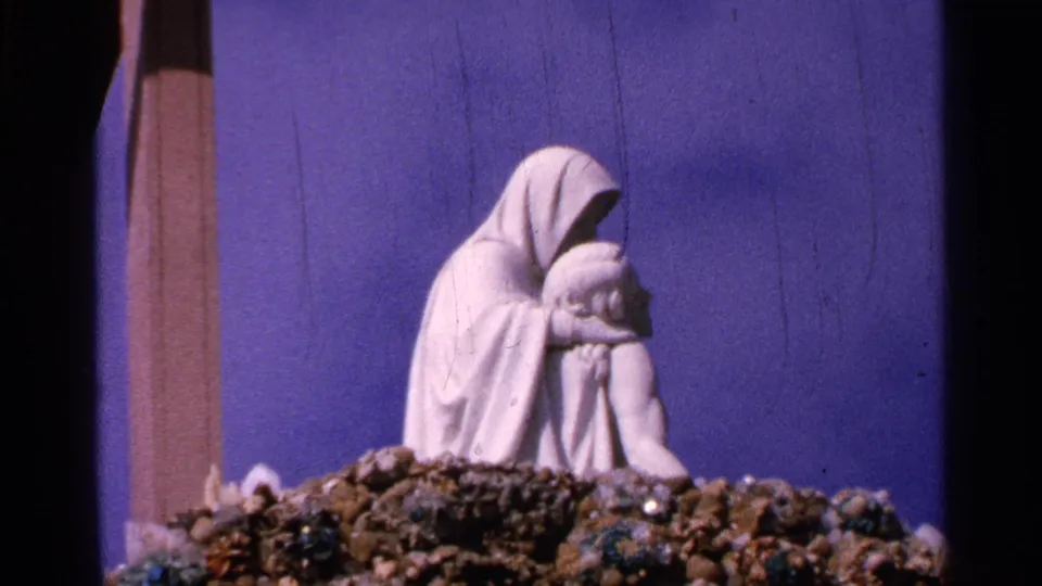 1965 Saint Louis frame showing the white Pieta-like sculpture and rocky base, with a dark divider and blue backdrop in view.