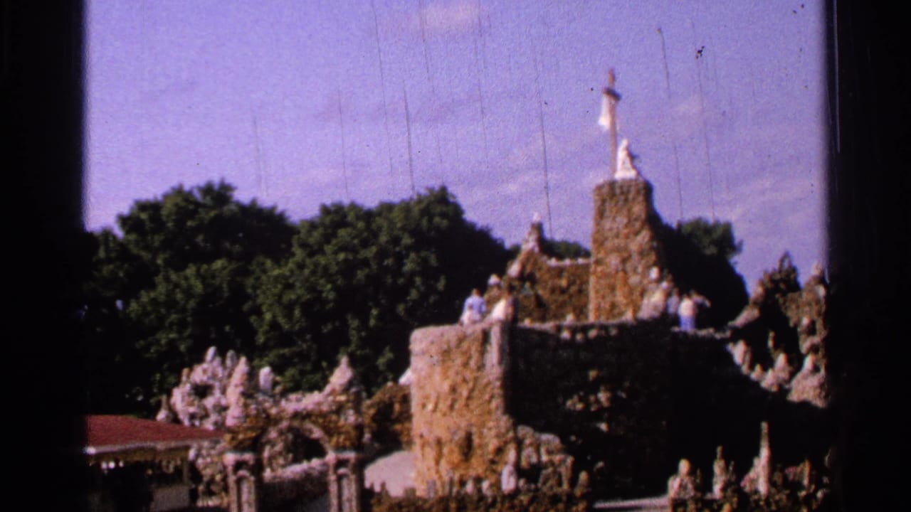 1965 Saint Louis 8mm frame showing visitors near an outdoor religious display with stone forms, statues, trees, and visible film wear.